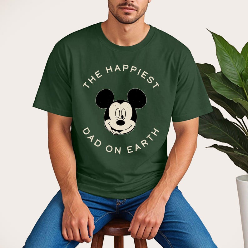 A man wearing a green t-shirt with a Mickey Mouse design and the text "The happiest dad on earth" is sitting on a stool.