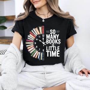 Può includere: T-shirt nero con un design a orologio fatto di libri. Le lancette dell'orologio segnano le 12. Il testo "SO MANY BOOKS SO LITTLE TIME" è stampato in bianco. La maglietta è girocollo.