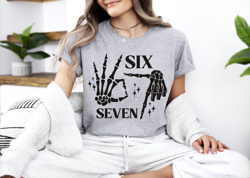 A woman wearing a grey t-shirt with a graphic design of a skeleton hand and the text "SIX SEVEN" is sitting on a couch.