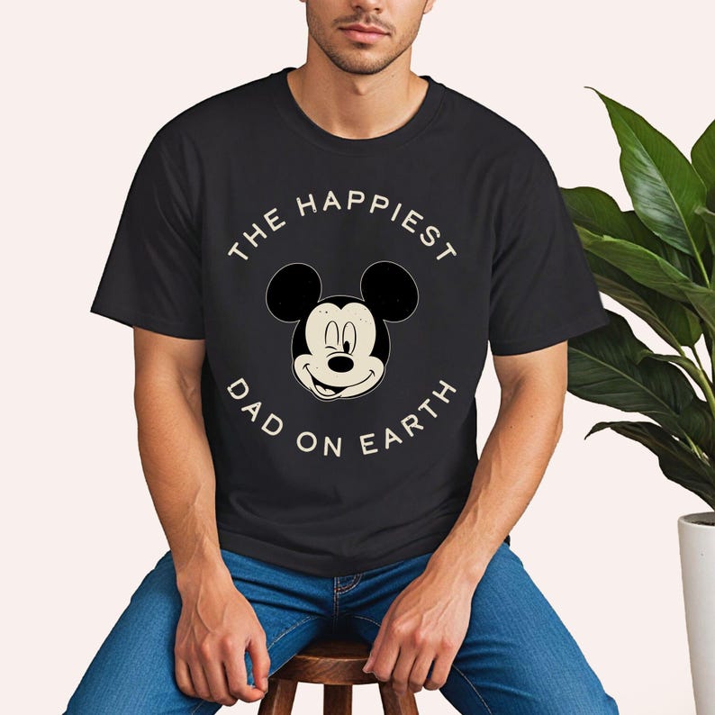 A man wearing a black t-shirt with a Mickey Mouse design and the text "The happiest dad on earth" is sitting on a stool in front of a potted plant.