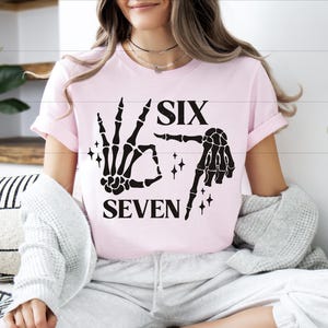 A woman wearing a pink t-shirt with a graphic design of a skeleton hand and the text "SIX SEVEN" is sitting on a couch.