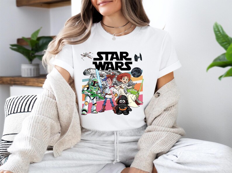 A woman wearing a white t-shirt with a Star Wars graphic and cartoon characters, including a character with a green alien head, is sitting in a room with plants and a striped pillow.