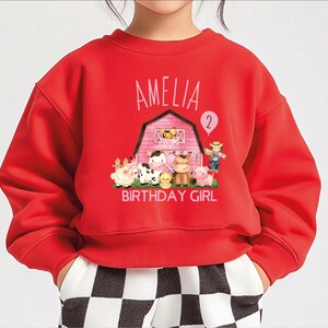 A red sweatshirt with a cartoon farm scene and the name "Amelia" printed on it, along with the number "2" and the text "BIRTHDAY GIRL".