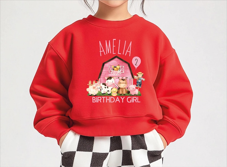 A red sweatshirt with a cartoon farm scene and the name "Amelia" printed on it, along with the number "2" and the text "BIRTHDAY GIRL".