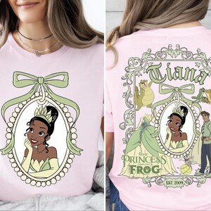 A woman wearing a pink t-shirt with a design featuring a cartoon image of a young girl, along with the text "Tiana the Princess Frog" and "Est. 2009".