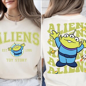 Puede incluir: Una camiseta blanca con un personaje extraterrestre verde de Toy Story. La parte delantera de la camiseta dice "Aliens Est. 1995 Toy Story". La parte trasera de la camiseta tiene un patr&oacute;n repetido del personaje extraterrestre y la palabra "Aliens" en una fuente retro.