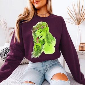 a woman sitting on a bed wearing a purple shirt with a green woman on it