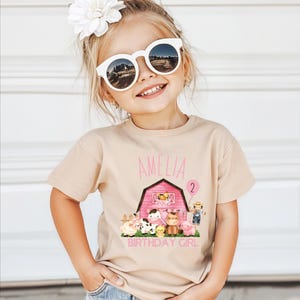 A young girl wearing a t-shirt with a farm-themed graphic and the name 'Amelia' printed on it, smiling and posing for a photo.