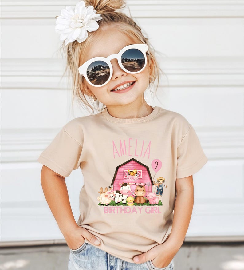 A young girl wearing a t-shirt with a farm-themed graphic and the name 'Amelia' printed on it, smiling and posing for a photo.