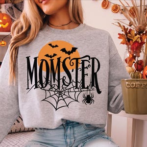 May include: A light grey crewneck sweatshirt featuring a black graphic print that reads MOMSTER in a stylised font. The design includes an orange full moon, three black flying bats, a spider web, and a small black spider hanging from the text.