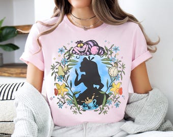 May include: Pink t-shirt with a graphic print of Alice in Wonderland. The graphic features Alice's silhouette, the Cheshire Cat, a pocket watch, and flowers.