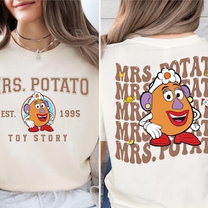 Può includere: T-shirt color crema con un design di Mrs. Potato Head di Toy Story. Il davanti mostra "MRS. POTATO" sopra l'illustrazione del personaggio, con "EST. 1995" e "TOY STORY" sotto. Il retro ripete "MRS. POTATO" con il personaggio.