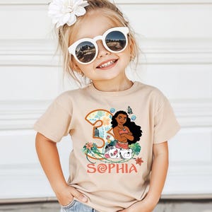 May include: A tan t-shirt featuring a cartoon illustration of a character with long black hair, a pink top, and a floral design. The number "3" and the name "Sophia" are also printed on the shirt. The child is wearing white sunglasses and a flower in her hair.