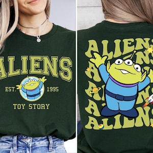 Puede incluir: Camiseta verde con un gr&aacute;fico de un extraterrestre de tres ojos de Toy Story. La parte delantera de la camiseta dice "Aliens Est. 1995 Toy Story". La parte trasera de la camiseta tiene un patr&oacute;n repetido del extraterrestre y la palabra "Aliens" en una fuente retro.