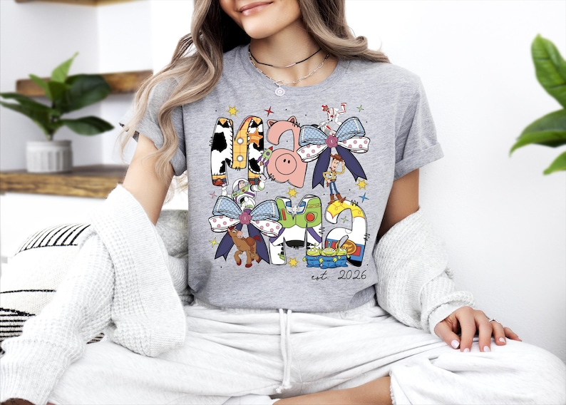 A woman wearing a gray t-shirt with a colorful graphic design and text, sitting on a couch with plants in the background.
