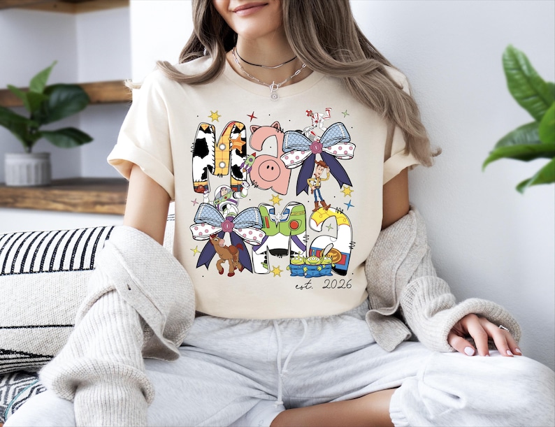A woman wearing a t-shirt with a cartoon character design and text, sitting on a couch with plants in the background.