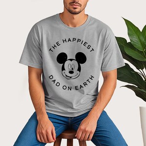 A man wearing a gray t-shirt with a Mickey Mouse design and the text "The happiest dad on earth" printed on it.