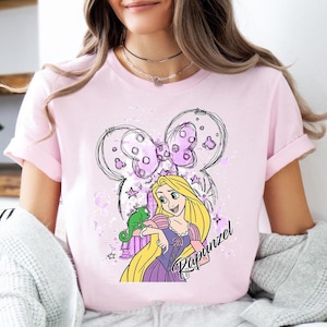 May include: A light pink t-shirt featuring a cartoon illustration of Rapunzel with long blonde hair, a green chameleon, and a purple dress. The design includes a Mickey Mouse-inspired bow and the name "Rapunzel".