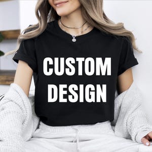 May include: Black t-shirt with the words "CUSTOM DESIGN" in large white block letters. The shirt has short sleeves and a crew neck. The model is wearing a light-colored sweater.