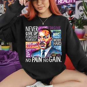 Peut inclure: Sweat-shirt noir avec un portrait coloré de Martin Luther King Jr. et des citations inspirantes. Le texte comprend des phrases comme « Never Give Up », « Dream Big » et « No Pain No Gain ».
