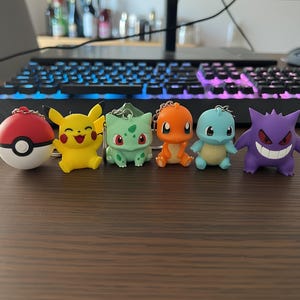 Op de afbeelding: Een set van zes kleine figuur-sleutelhangers met een rood-witte Poké Ball, een gele Pikachu, een lichtgroene Bulbasaur, een oranje Charmander, een lichtblauwe Squirtle en een paarse Gengar, elk voorzien van een zilverkleurige metalen ring.