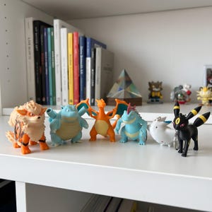 Puede incluir: Una colección de seis llaveros de plástico con diversos personajes, incluyendo un cánido naranja y crema, una criatura azul similar a una tortuga, un dragón naranja, un personaje bípedo azul, una figura blanca redondeada y un personaje negro tipo zorro.