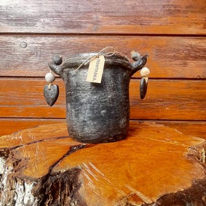 Peut inclure: Pot en céramique noire avec deux anses et une étiquette indiquant "MONTANEA". Le pot est décoré de petites perles blanches et grises suspendues aux anses.