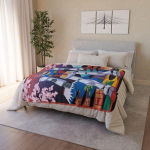 Puede incluir: Una colorida manta con una ilustración detallada de un robot, sobre un fondo de flores de cerezo y paisajes urbanos. El diseño incorpora tonos de azul, rojo, amarillo y blanco, con un borde naranja. La manta se muestra en una cama con ropa de cama blanca.