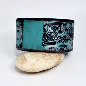 May include: A black and turquoise polymer clay bracelet with a swirling pattern. The bracelet is sitting on a light brown rock.