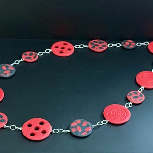May include: A necklace featuring red and black circular beads connected by a silver chain. The red beads have a variety of designs, including concentric circles and dot patterns. The necklace is displayed against a black background.
