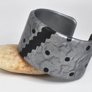 May include: A silver and black patterned cuff bracelet with a textured design. The bracelet has a wide band and is made of polymer clay.