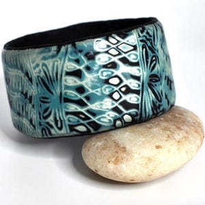 May include: A wide, teal blue and black patterned polymer clay bangle bracelet. The bracelet has a geometric design with white accents.