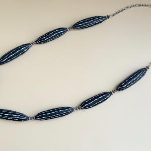 May include: A necklace featuring elongated, dark blue beads with white striped patterns. The beads are connected by small silver-toned metal links and a delicate chain. The necklace is displayed on a light beige surface.
