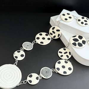 May include: A black and white necklace with a variety of circular pendant designs, including polka dots, swirls, and abstract shapes. The necklace is made of white ceramic and silver chain.
