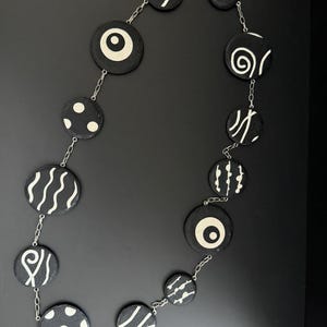 May include: A black and white necklace with round wooden beads. Each bead is painted with a different geometric design, including waves, dots, spirals, and lines.