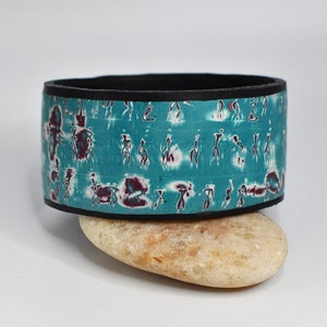 May include: A teal ceramic bowl with a black rim and a white and purple abstract design. The bowl is sitting on a light brown rock.