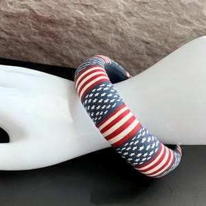 May include: A fabric-covered bangle bracelet featuring a patriotic design. The bracelet displays alternating red and white stripes with a blue field of white stars, reminiscent of the American flag. The bangle is a circular shape.