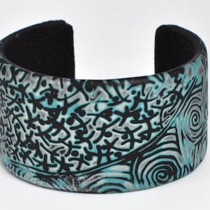 May include: A wide black and teal blue polymer clay cuff bracelet with a swirling, abstract pattern.