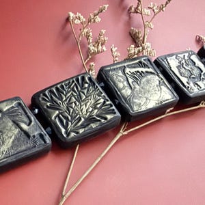 May include: A black bracelet with gold accents features five square tiles with different embossed designs. The tiles are connected by a thin gold chain.
