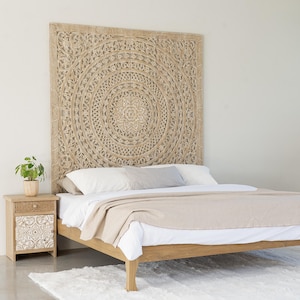 May include: A wooden bed frame with a carved headboard. The headboard has a circular pattern. The bed is made with white sheets and a beige blanket. A small bedside table with a drawer is on the left. The bed frame is approximately 200 cm long.