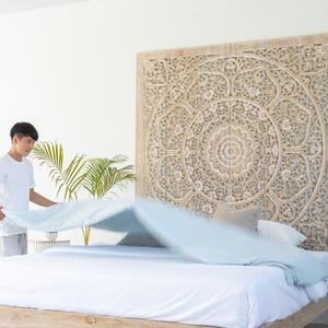 May include: A white bed with a light blue blanket being pulled up by a person. The headboard is a large, intricately carved wooden panel with a circular design.