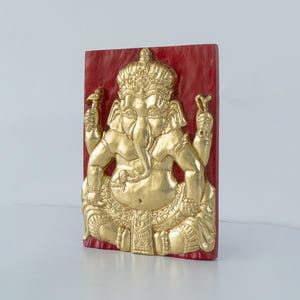Op de afbeelding: Een goudkleurige reliëfsculptuur van de hindoeïstische godheid Ganesha, zittend met meerdere armen, op een rechthoekige rode achtergrond. De sculptuur heeft ingewikkelde details, waaronder een kroon en sierlijke kleding. Het kunstwerk wordt gepresenteerd tegen een effen witte achtergrond.