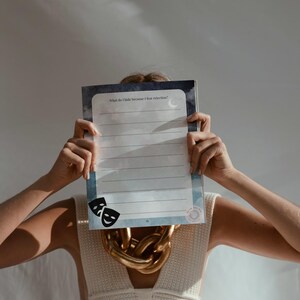 Puede incluir: Un diario con el texto "What do I hide because I fear rejection?" se sostiene. El diario tiene un diseño de acuarela azul y blanco con una luna y dos máscaras de teatro. Se ve un collar de cadena dorada.