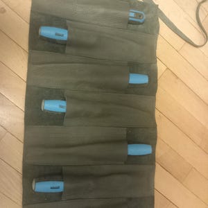 Peut inclure: Un rouleau à outils en cuir vert foncé avec plusieurs poches, chacune contenant un outil à manche bleu. Le rouleau est doté d'une sangle de transport et est conçu pour ranger et protéger les outils. Le cuir a un aspect texturé.
