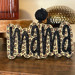 May include: A decorative sign featuring the word mama in black glitter script lettering layered over a tan and brown leopard print background. The piece includes a black cord loop at the top for hanging, with a small black glittery pom-pom attached.