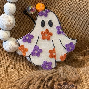 May include: A white glittery ghost-shaped Halloween decoration with black eyes and orange and purple flower accents. The ghost is hanging with a black string. Decorative wooden beads and a small wooden block are also visible.