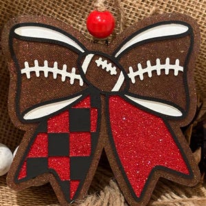 May include: A decorative bow-shaped ornament featuring a brown glitter finish, a central football graphic, and two dangling tails. One tail has a red and black checkered pattern, while the other is solid red glitter. A red bead sits above the bow.