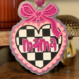 May include: A heart-shaped hanging ornament featuring a black and white checkered pattern with the word mama in pink glittery script. The ornament is framed by layers of pink and silver glitter, topped with a pink glitter bow and a pink bead.