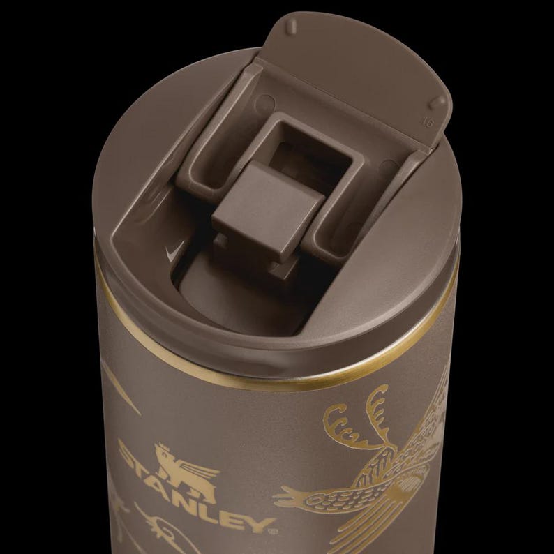 Puede incluir: Taza de viaje Stanley marr&oacute;n con tapa abatible. La taza presenta elementos decorativos dorados y el logotipo de Stanley. La tapa est&aacute; abierta, revelando el interior. La taza est&aacute; dise&ntilde;ada para bebidas fr&iacute;as o calientes.