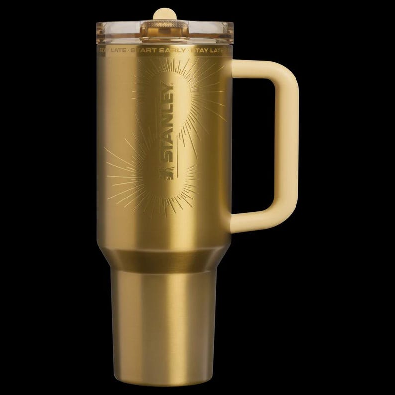 May include: A gold-colored Stanley travel mug with a cream-colored handle and lid. The mug features the Stanley logo and a sunburst design. The mug is designed to keep beverages hot or cold.